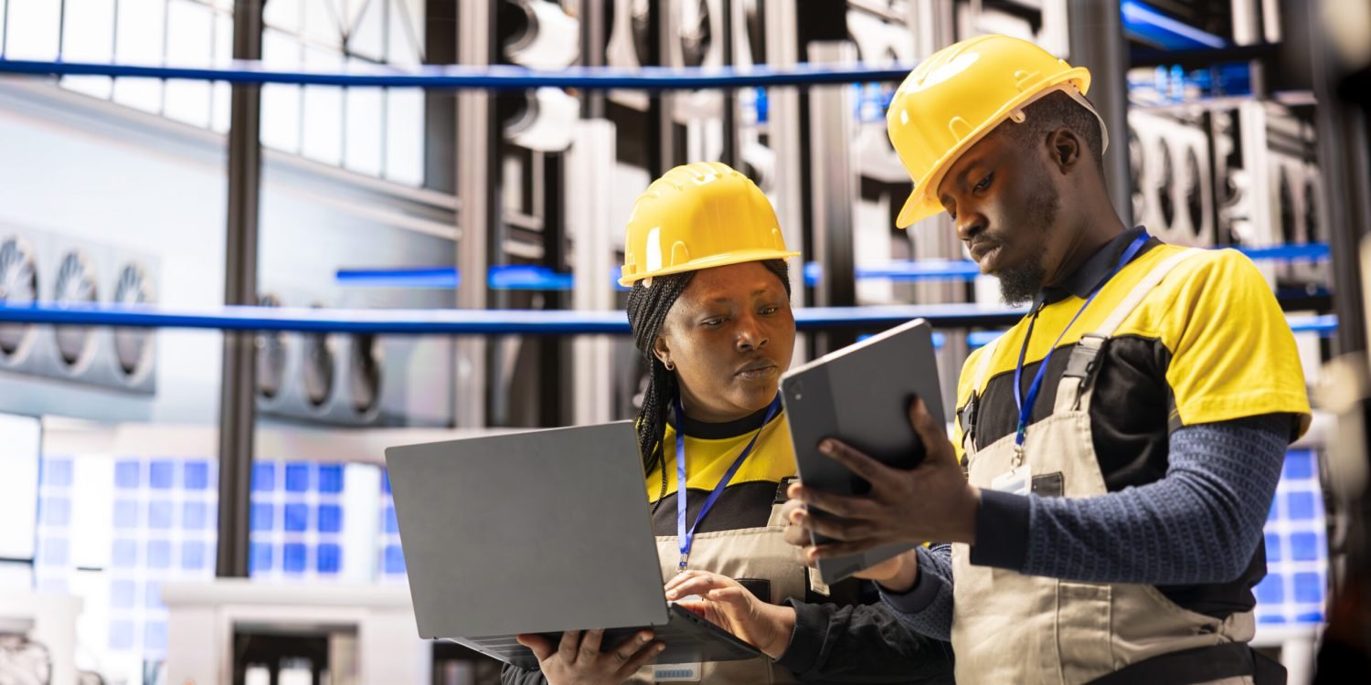 African american engineering team with laptop and tablet, collecting maintenance data in automated factory. Professional technicians using industry 4.0 software in smart manufacturing power plant.